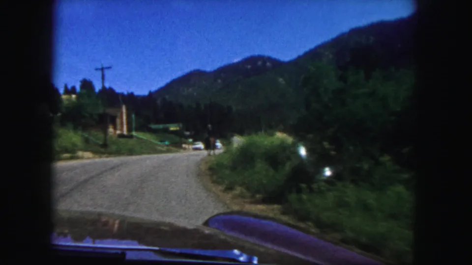 Passenger-seat view on a paved mountain road near Colorado Springs, with a car hood, roadside brush, utility pole, and wooded hills.