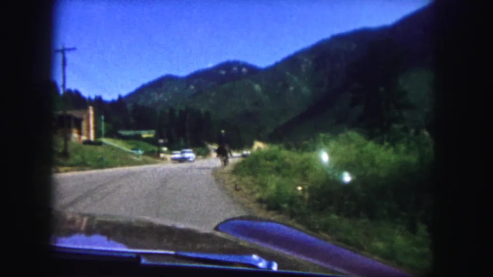 1958 home-movie road view showing an oncoming automobile on a paved mountain road, with trees, brush, and hills beyond.