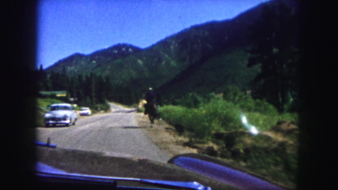 1958 Colorado Springs home-movie frame showing a paved mountain road, classic cars, and a horseback rider in the distance.