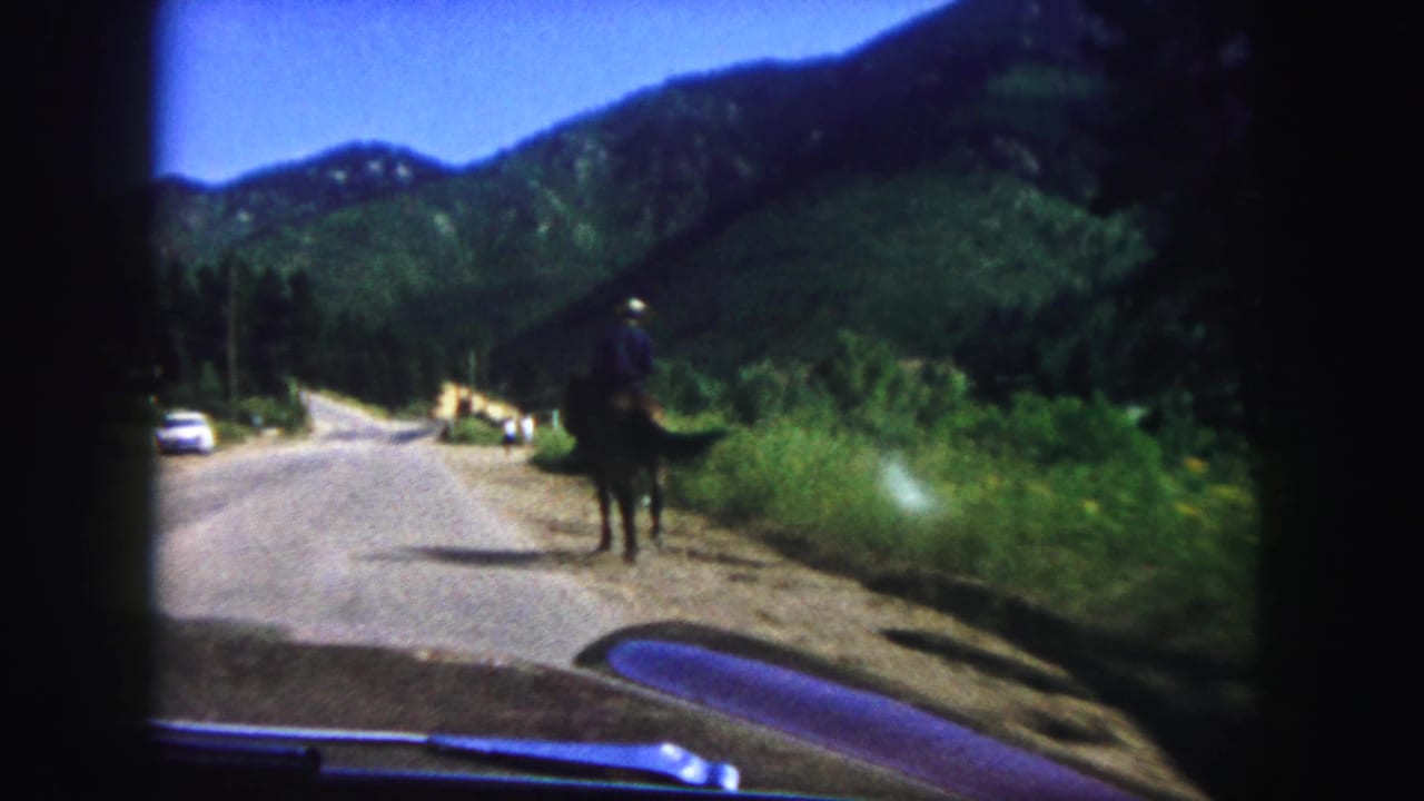 Horseback rider travels along the roadside beside a mountain road and parked or passing cars.