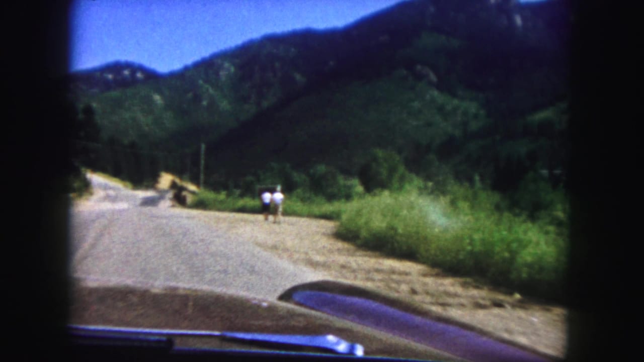 1958 Colorado Springs home-movie view from inside a car approaching two pedestrians on a mountain road beneath green hills.