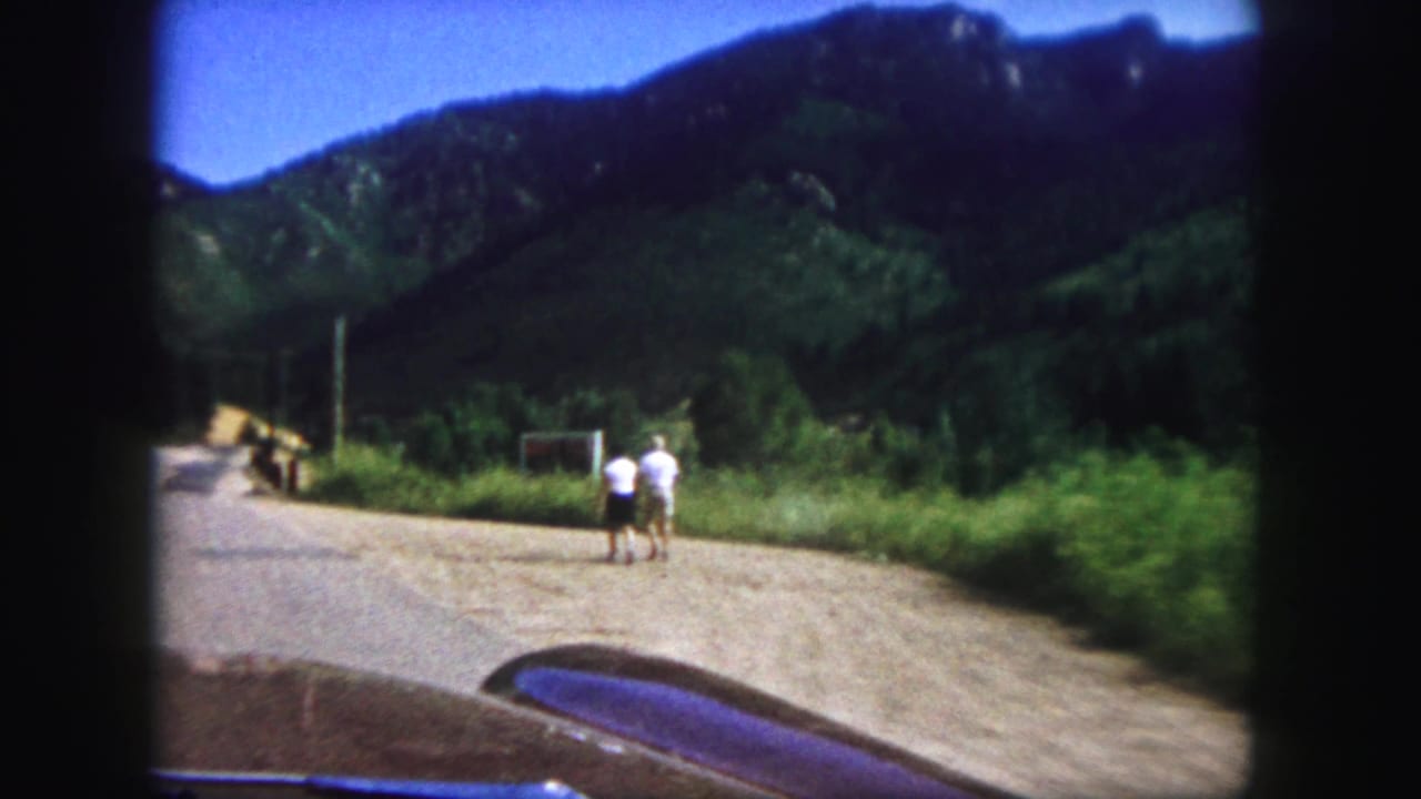 Mountain road seen from a car as two pedestrians walk near a roadside sign or structure.