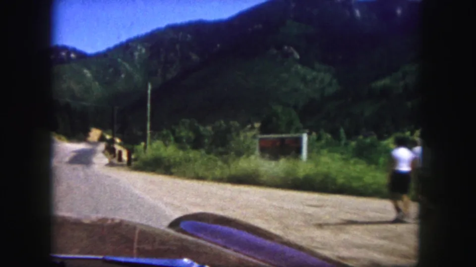 The vehicle passes two roadside pedestrians in a 1958 Colorado Springs mountain travel scene, with road, brush, and utility pole visible.