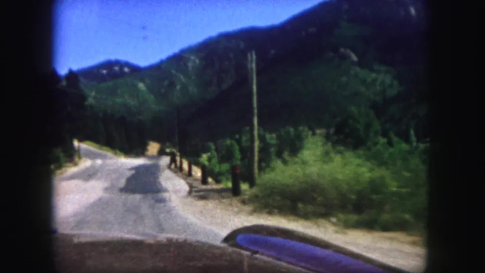 Passenger-view 8mm footage on a Colorado Springs mountain road after passing pedestrians, with a road edge, posts, and hills ahead.