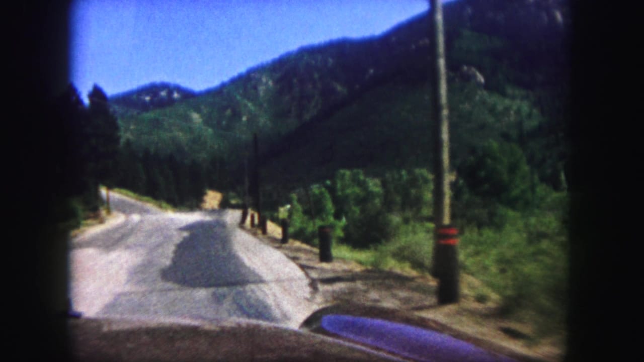 Car windshield view of a curving mountain road bordered by pine forest and hills.