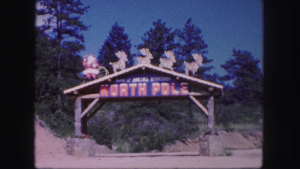 1958 Colorado Springs home-movie frame showing a wooden North Pole archway topped with Santa and reindeer figures among pine trees.