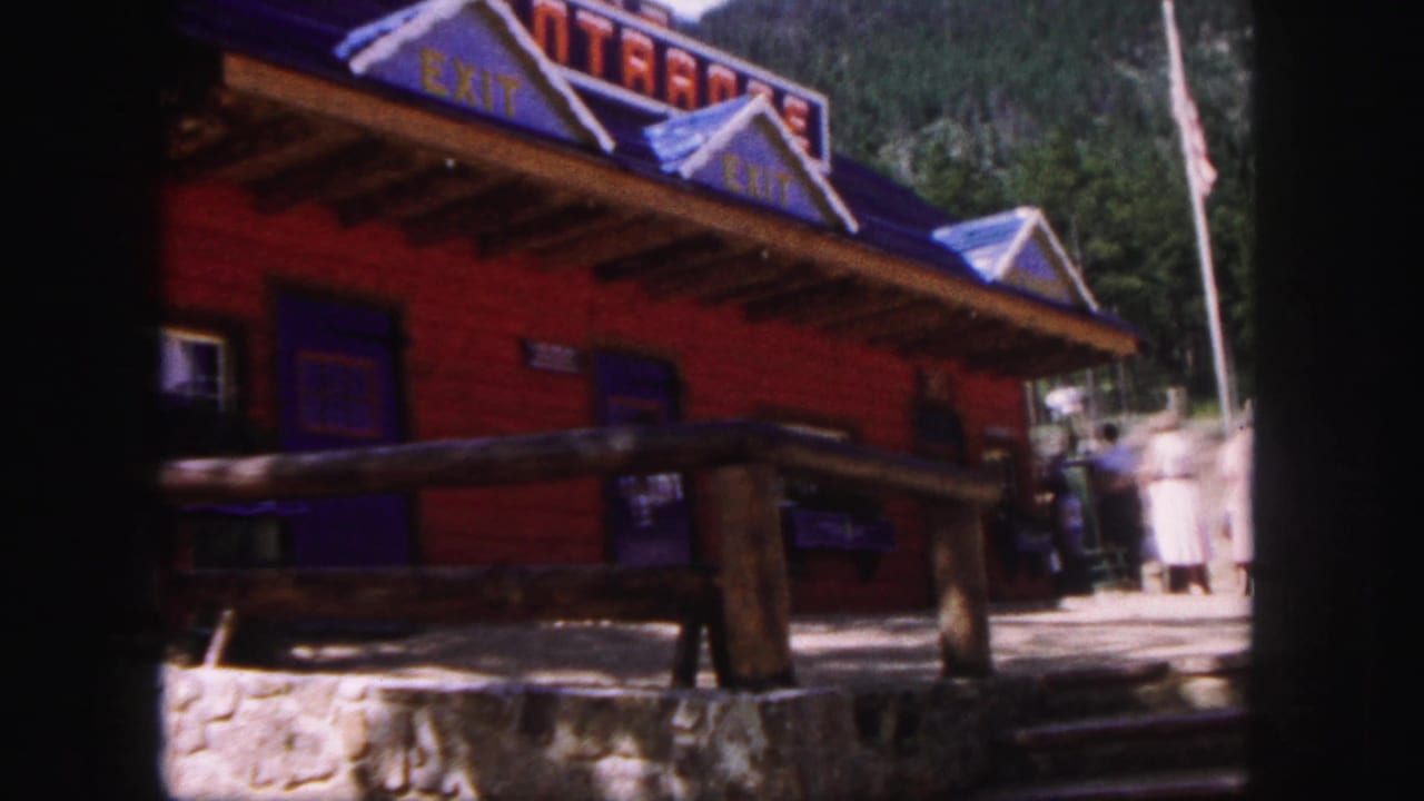 1958 Colorado Springs 8mm frame of a red log-style roadside building with blue roof trim, exit lettering, railing, stonework, and visitors nearby.