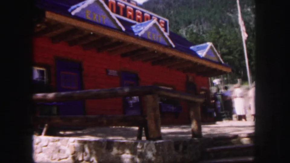 1958 Colorado Springs 8mm frame of a red log-style roadside building with blue roof trim, exit lettering, railing, stonework, and visitors nearby.