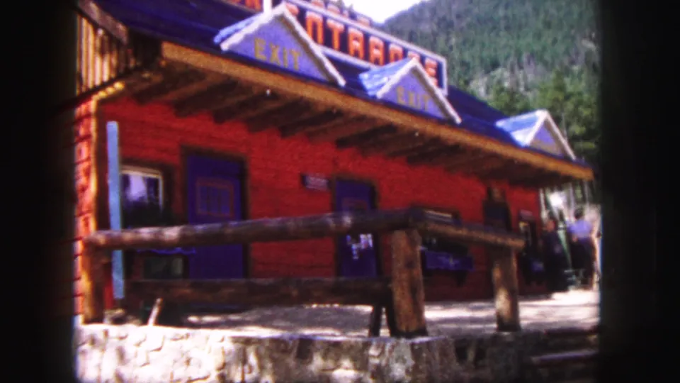 1958 Colorado Springs 8mm view of the red rustic exterior with blue trim, log porch railing, stone base, wooded background, and people by the entrance.