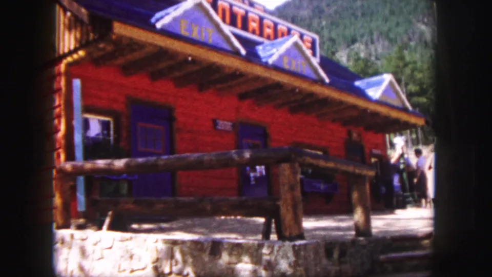 1958 Colorado Springs home-movie frame of a red log-style building with blue roof trim, visible exit lettering, log railing, stone foundation, and visitors nearby.