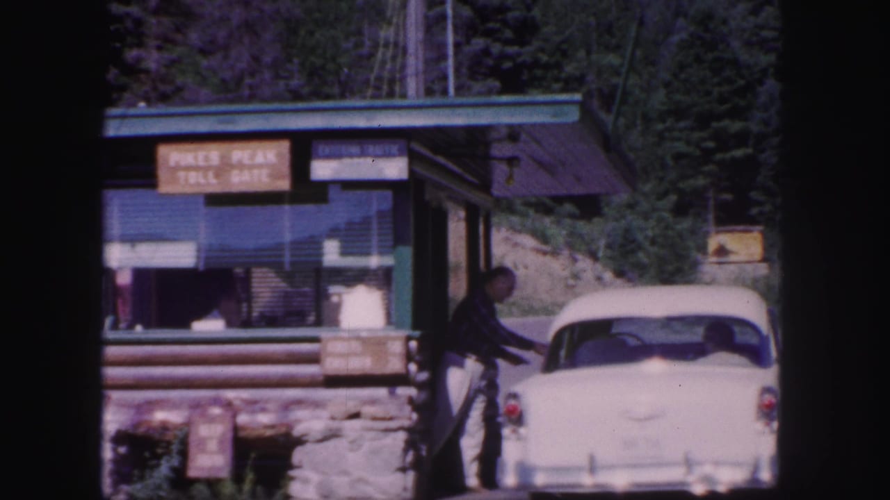 1958 Colorado Springs footage showing a white car at the Pikes Peak Toll Gate, with a man near the vehicle and pine trees behind the booth.