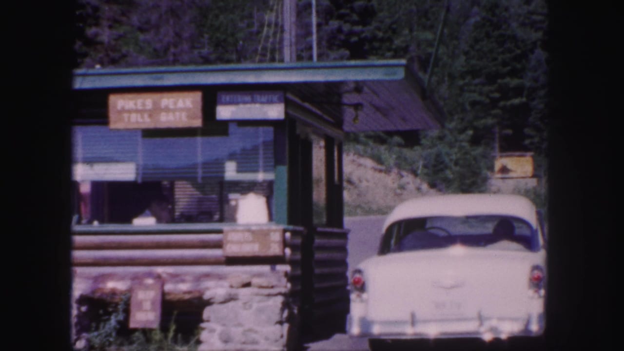 White car at the toll booth, with Pikes Peak Toll Gate signage and pine-covered hillside behind it.