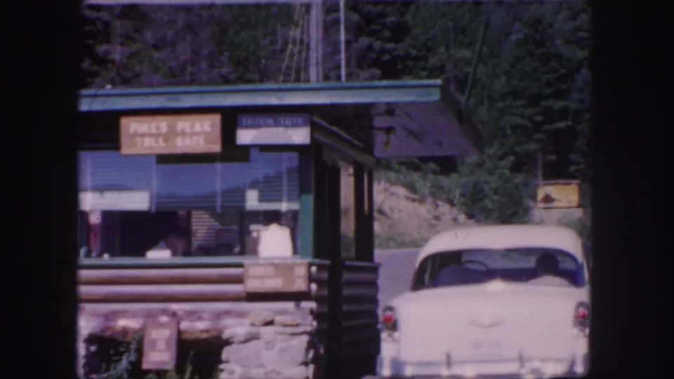 Final frame of the 1958 Pikes Peak Toll Gate scene, with the white car, booth signage, log-and-stone structure, and forested roadside visible.