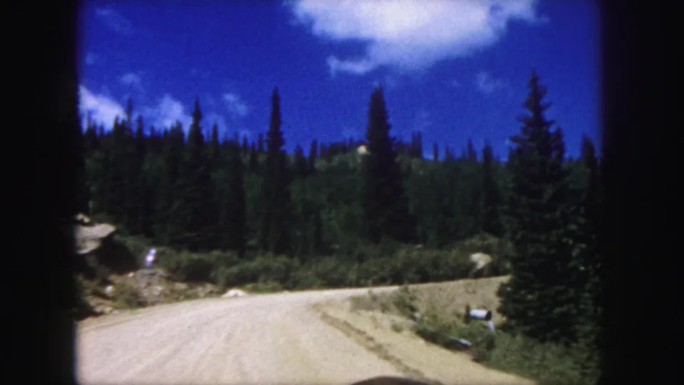 1958 travel footage view from a car on a pale dirt road running past forested slopes near Colorado Springs.