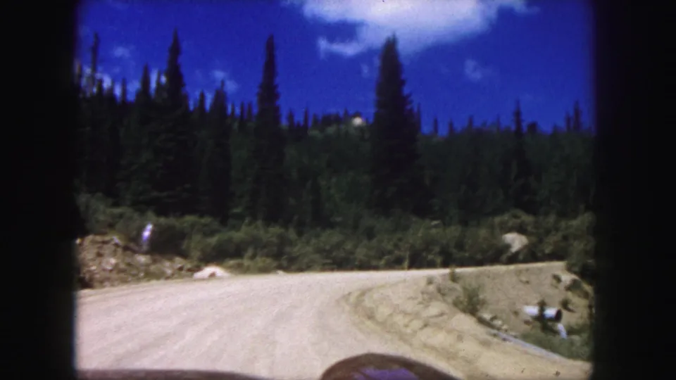 In-car home-movie frame showing a curving dirt road, roadside rocks, and pine forest in the Pikes Peak area.