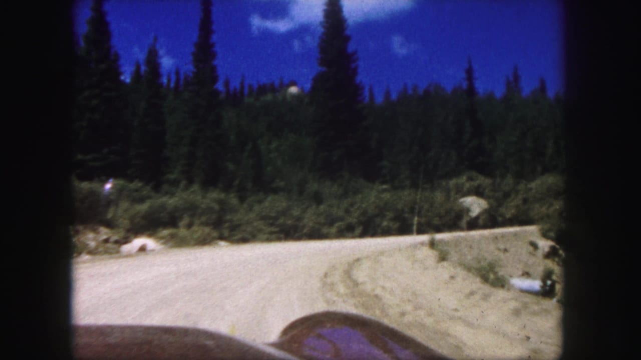 1958 Colorado Springs 8mm frame showing a car approaching a pale dirt mountain road bordered by dense evergreens and blue sky.