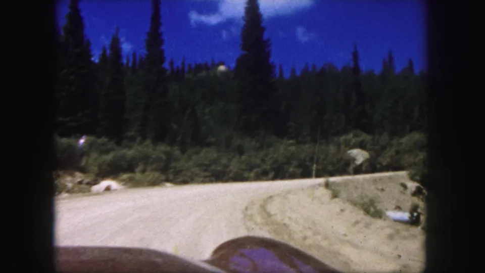 1958 Colorado Springs 8mm frame showing a car approaching a pale dirt mountain road bordered by dense evergreens and blue sky.