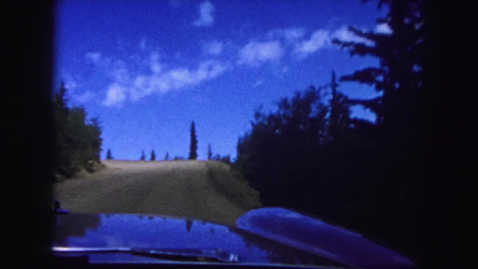 1958 8mm home-movie frame from inside a car on a Colorado Springs mountain road, with hood reflection and trees along the slope.