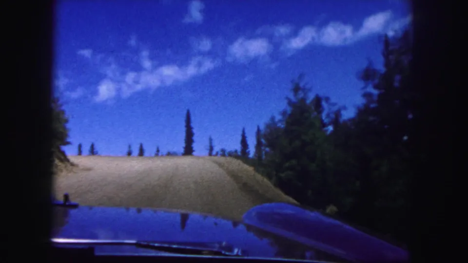 Colorado Springs 1958 8mm frame showing the car nearing a hilltop on a pale dirt road bordered by evergreens beneath scattered clouds.