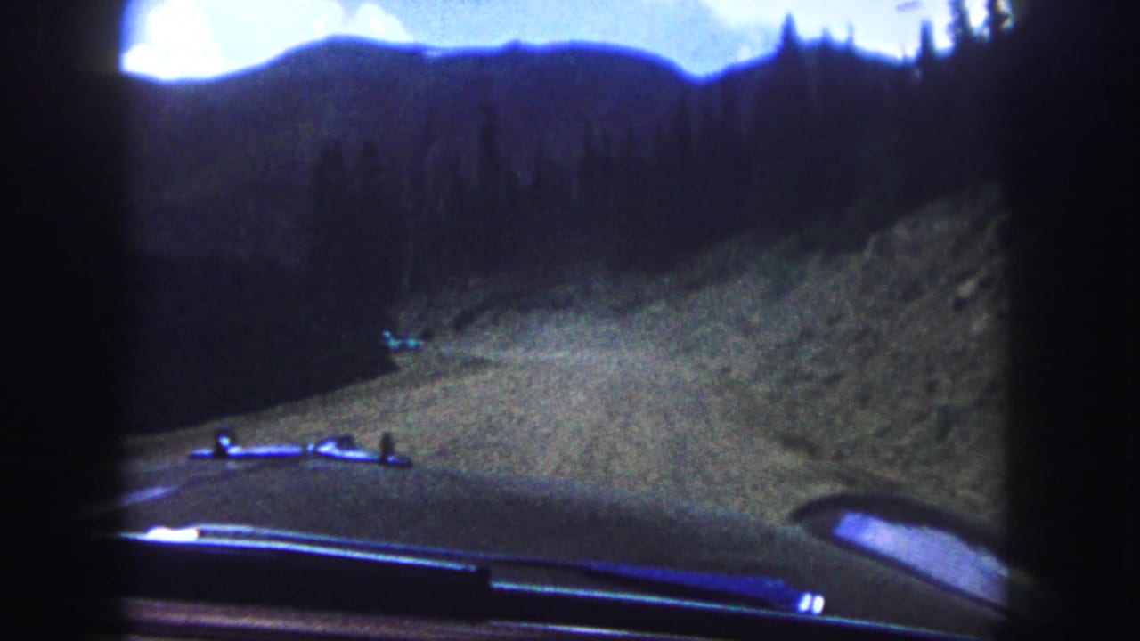 Car hood and windshield wipers face a curving hillside road with dark pine forest and mountains beyond.