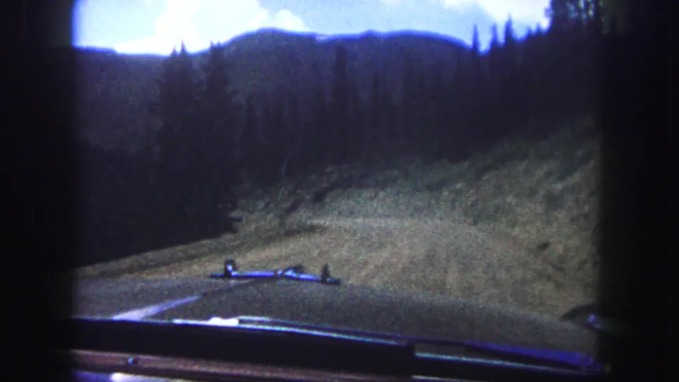1958 Colorado Springs frame from inside a moving car, looking toward a winding dirt or gravel mountain road through pine forest.