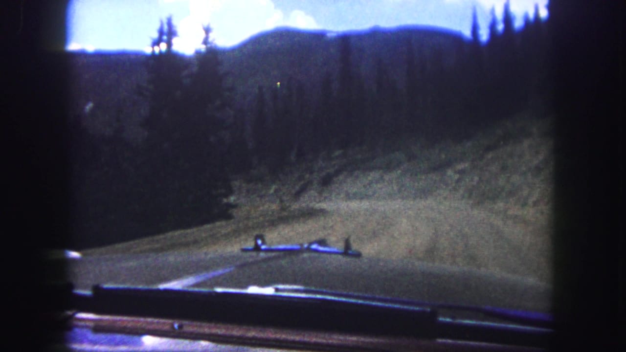1958 8mm view from inside a car on a paved mountain road near Colorado Springs, with pine hills and sky ahead.