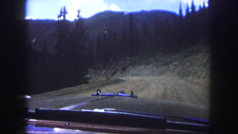 1958 8mm view from inside a car on a paved mountain road near Colorado Springs, with pine hills and sky ahead.