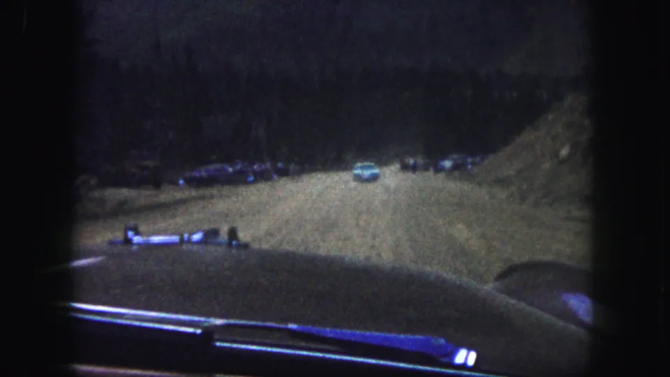 1958 home-movie frame from a moving car, with a light blue vehicle and parked cars along a wooded mountain road.