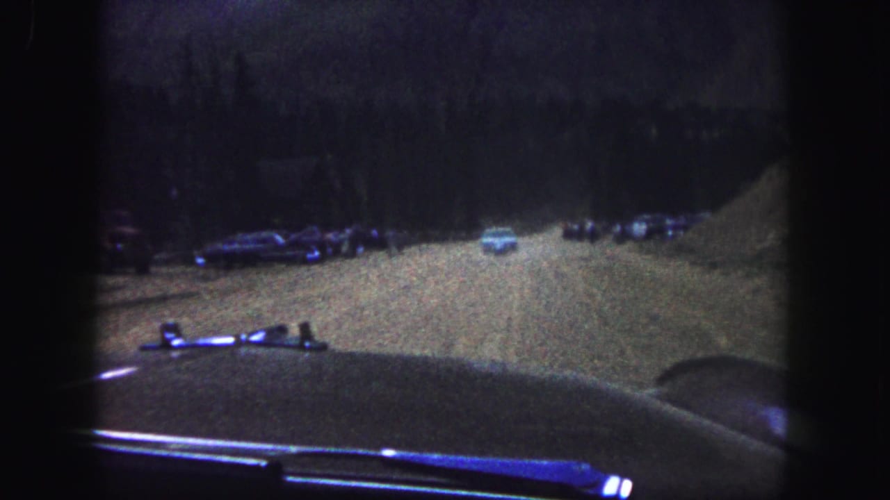 1958 Colorado Springs home-movie frame showing a dim dirt road from inside a car, with parked vehicles and dark trees ahead.