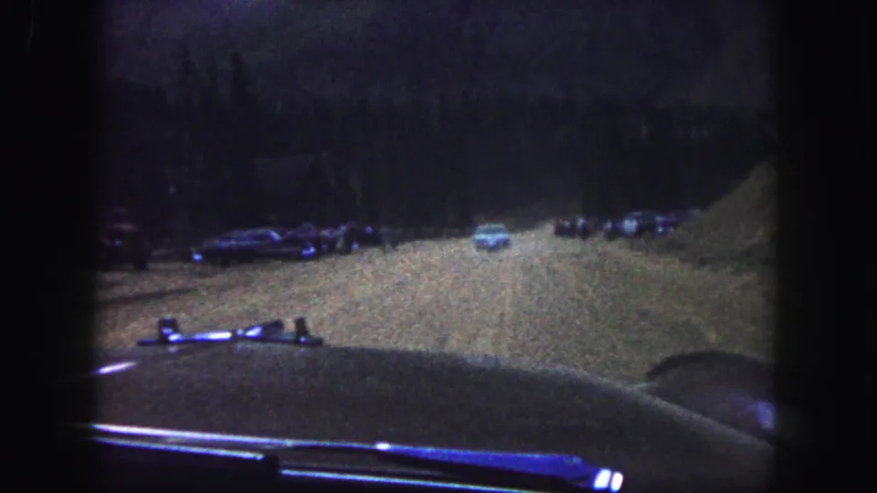1958 Colorado Springs home-movie frame showing a dim dirt road from inside a car, with parked vehicles and dark trees ahead.