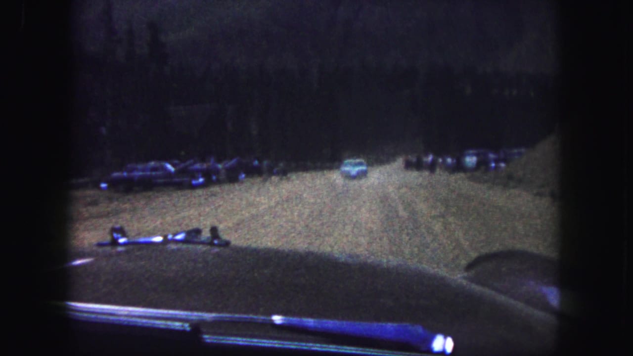 Dim windshield view of a dirt road with parked vehicles and a pale car ahead.