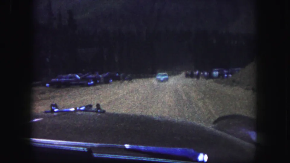 From inside a car in Colorado Springs, the frame shows a low-light 8mm view of a dirt road, parked cars, and a pale car farther ahead.