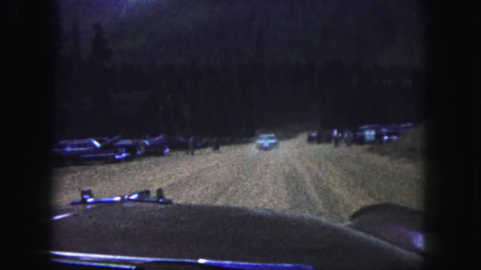 1958 Colorado Springs frame looking through a windshield toward a dirt road, parked cars, trees, and small distant people near the roadside.