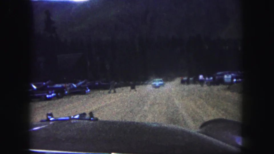 Colorado Springs home-movie frame from 1958 showing a windshield view of pedestrians crossing or standing near a dim dirt road with parked cars.
