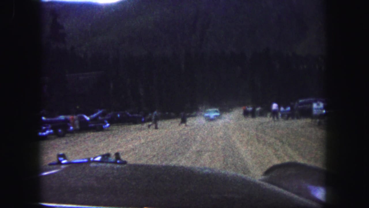 1958 Colorado Springs home-movie frame showing parked cars and distant people in a dark outdoor dirt clearing.