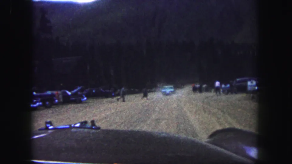 1958 Colorado Springs home-movie frame showing parked cars and distant people in a dark outdoor dirt clearing.