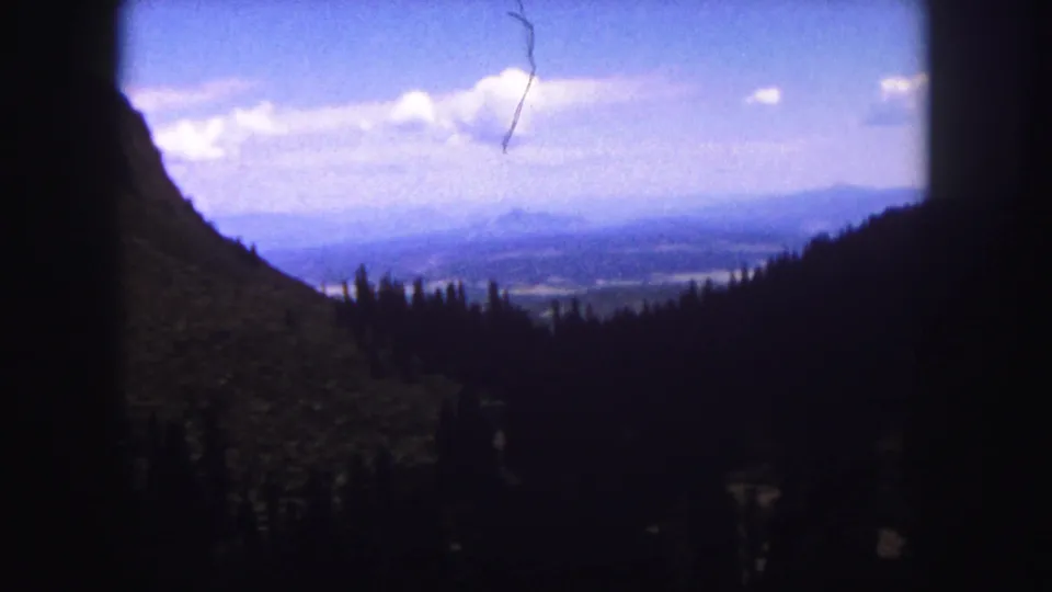 Handheld 1958 Colorado mountain view with dark trees, sloping terrain, distant valley, and visible film scratch.