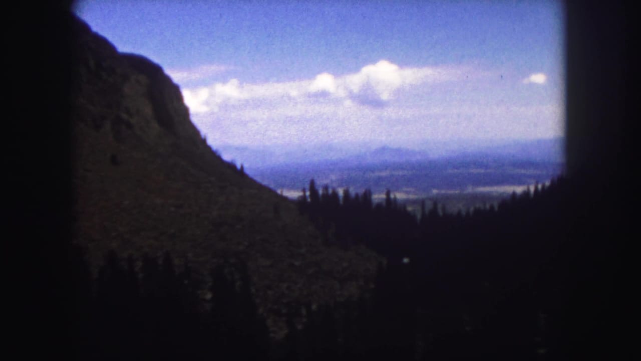1958 Colorado Springs home-movie frame showing a rocky mountainside, shadowed evergreens, distant valley, and bright clouded sky.