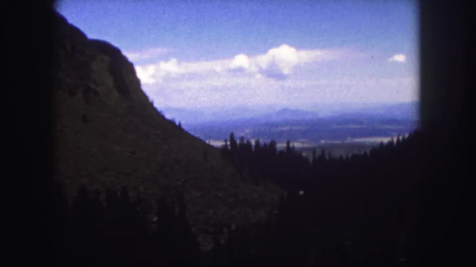 1958 Colorado Springs home-movie frame showing a rocky mountainside, shadowed evergreens, distant valley, and bright clouded sky.