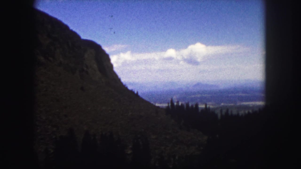 Mountain shoulder and distant valley seen through dark edge framing beneath a clouded blue sky.
