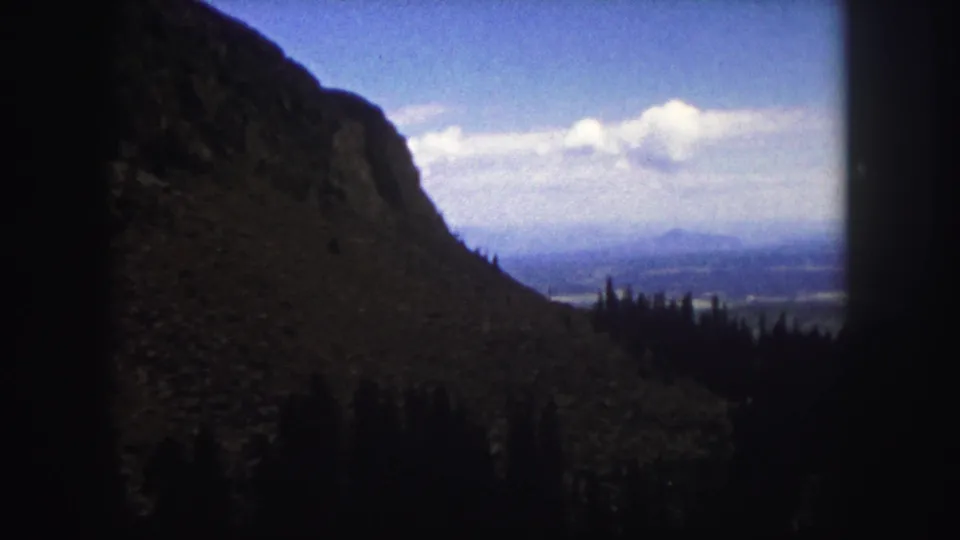 1958 Colorado Springs home-movie still showing a dark foreground slope, evergreen ridge, hazy distance, and bright cloud bank.