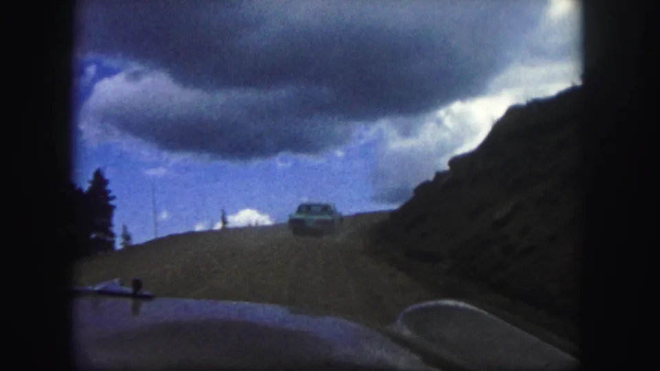 1958 Colorado Springs road-trip frame showing a dirt road between slopes, a distant car, heavy clouds, and a partial vehicle hood.