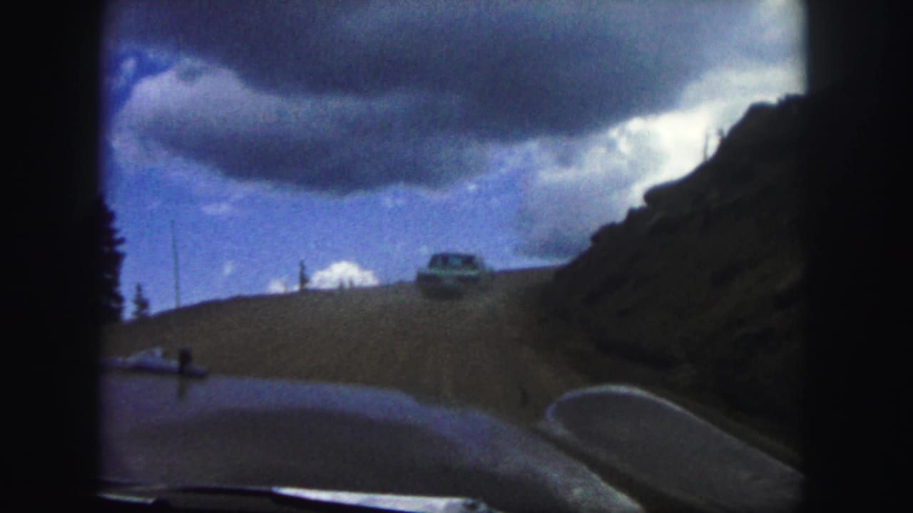 1958 Colorado Springs home-movie view through a car windshield, showing a dirt road, rocky slope, and another car ahead under heavy clouds.