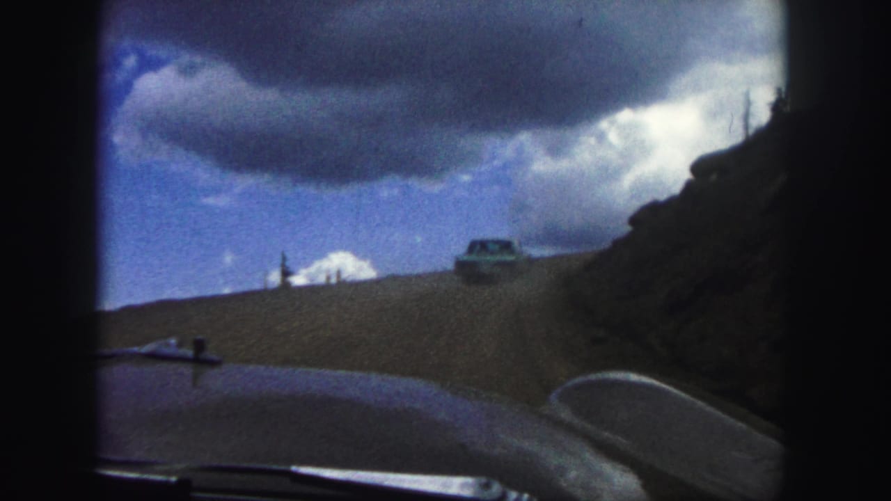 Car ahead on a curving dirt road seen from inside another vehicle.