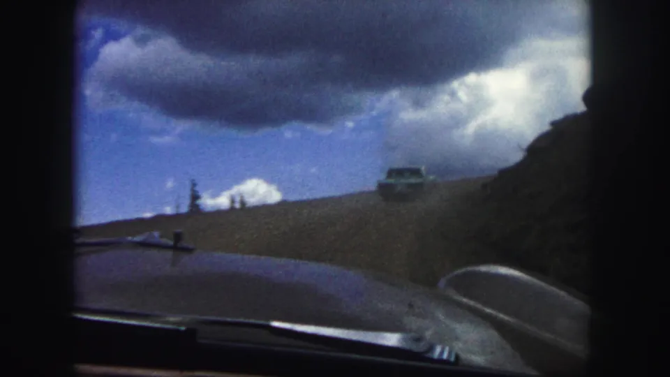 Home-movie frame from a 1958 Colorado Springs drive, with dashboard foreground, roadside pine, dirt road, and car ahead in mountain scenery.