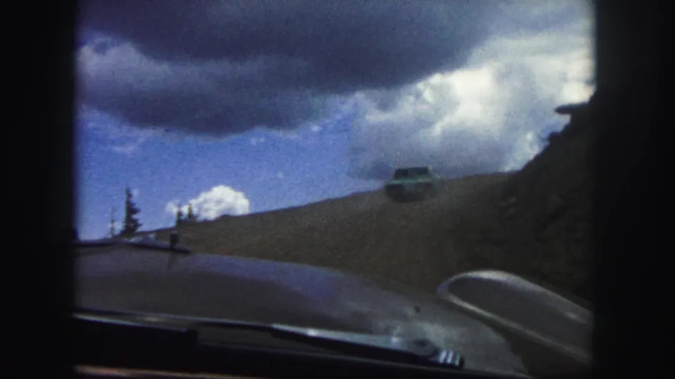 Colorado Springs road-trip frame from 1958 showing a windshield-framed dirt road, distant car, rocky hillside, pine tree, and dramatic cloud cover.