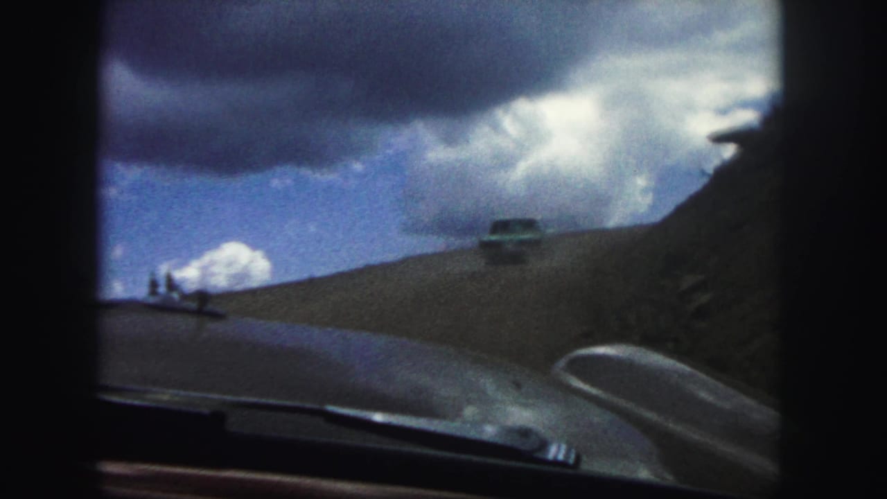 Car ahead on a dirt mountain road seen over the hood of a moving vehicle.