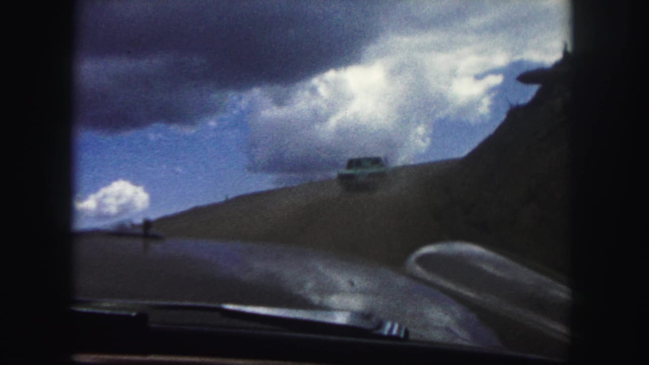 Distant car on a curving dirt road with vehicle hood visible in foreground.