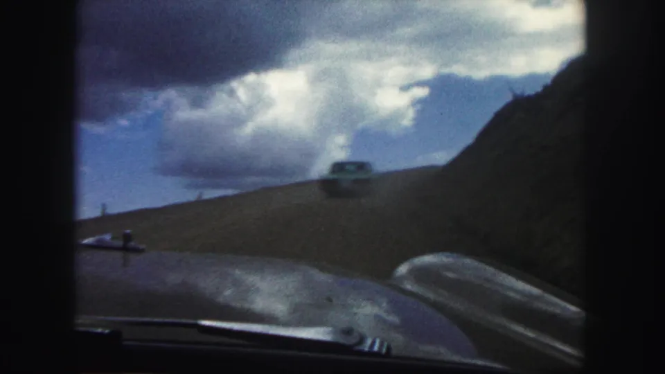 A 1958 in-car view shows a car ahead on an unpaved road in the Colorado Springs area, with dark film borders and bright clouds overhead.