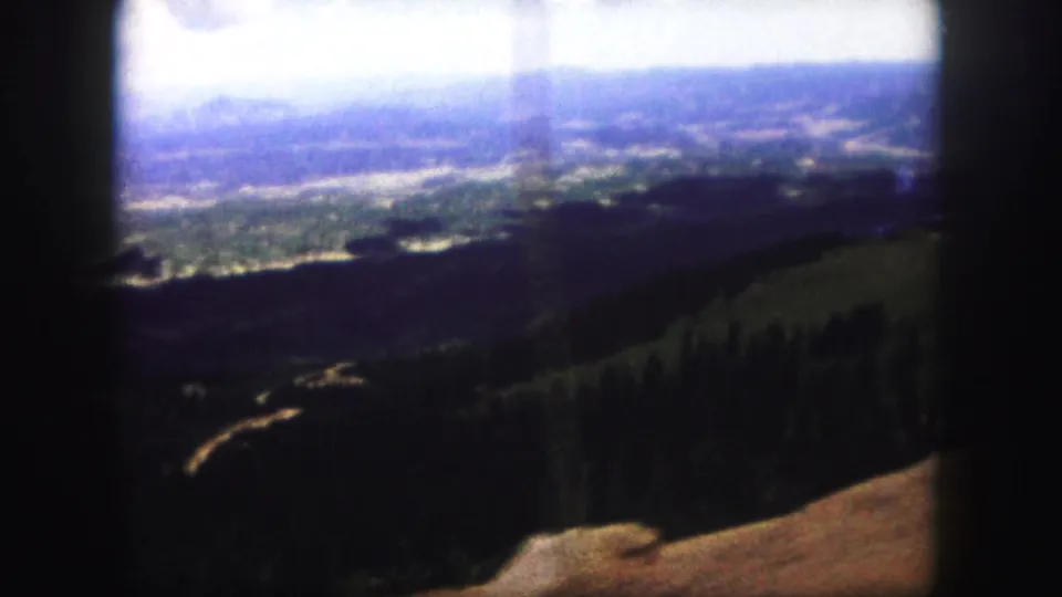 1958 8mm landscape frame with a hazy valley beyond dark woods and a pale winding road on the hillside.